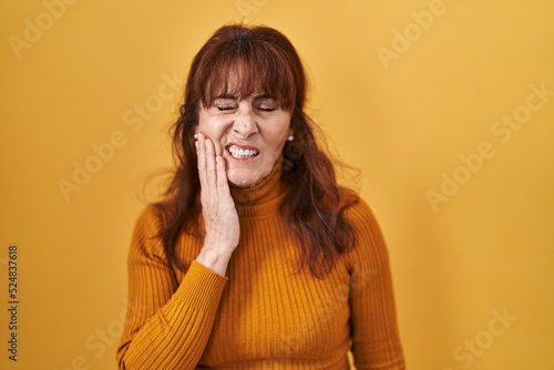 Middle age hispanic woman standing over yellow background touching mouth with hand with painful expression because of toothache or dental illness on teeth. dentist