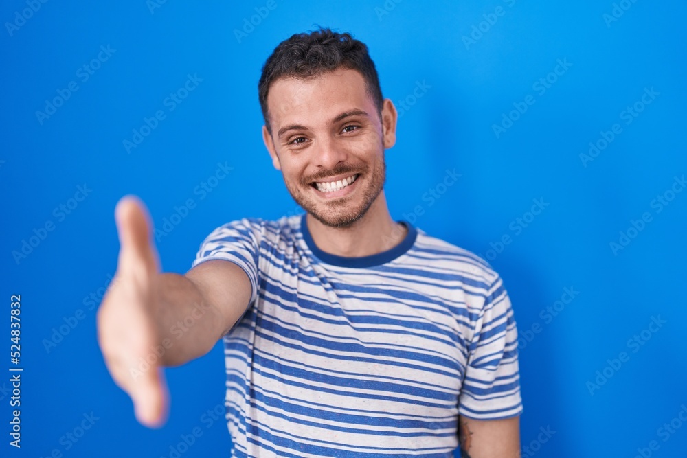 Young hispanic man standing over blue background smiling friendly offering handshake as greeting and welcoming. successful business. Young hispanic man standing over blue background smiling friendly offering handshake as greeting and welcoming. successful business.