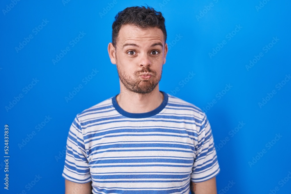 Young hispanic man standing over blue background puffing cheeks with funny face. mouth inflated with air, crazy expression. Young hispanic man standing over blue background puffing cheeks with funny face. mouth inflated with air, crazy expression.