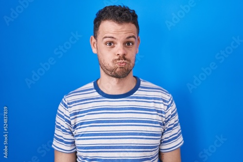 Young hispanic man standing over blue background puffing cheeks with funny face. mouth inflated with air, crazy expression.
