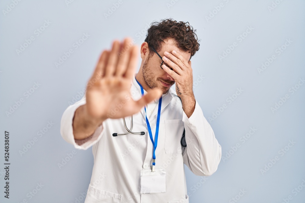 Young hispanic man wearing doctor uniform and stethoscope covering eyes with hands and doing stop gesture with sad and fear expression. embarrassed and negative concept. Young hispanic man wearing doctor uniform and stethoscope covering eyes with hands and doing stop gesture with sad and fear expression. embarrassed and negative concept.