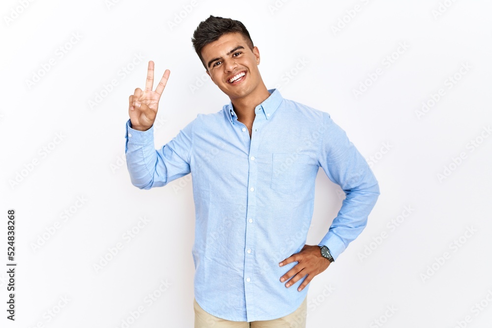 Young hispanic man wearing business shirt standing over isolated background smiling looking to the camera showing fingers doing victory sign. number two. Young hispanic man wearing business shirt standing over isolated background smiling looking to the camera showing fingers doing victory sign. number two.