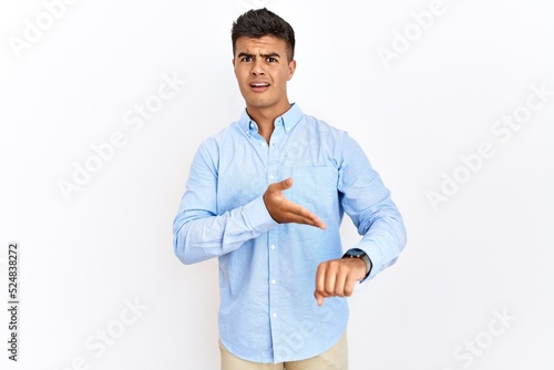 Young hispanic man wearing business shirt standing over isolated background in hurry pointing to watch time, impatience, upset and angry for deadline delay