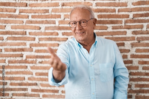 Senior man with grey hair standing over bricks wall smiling cheerful offering palm hand giving assistance and acceptance.