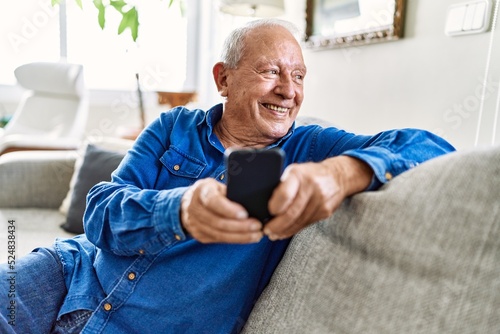 Senior man with grey hair sitting on the sofa at the living room of his house using smartphone