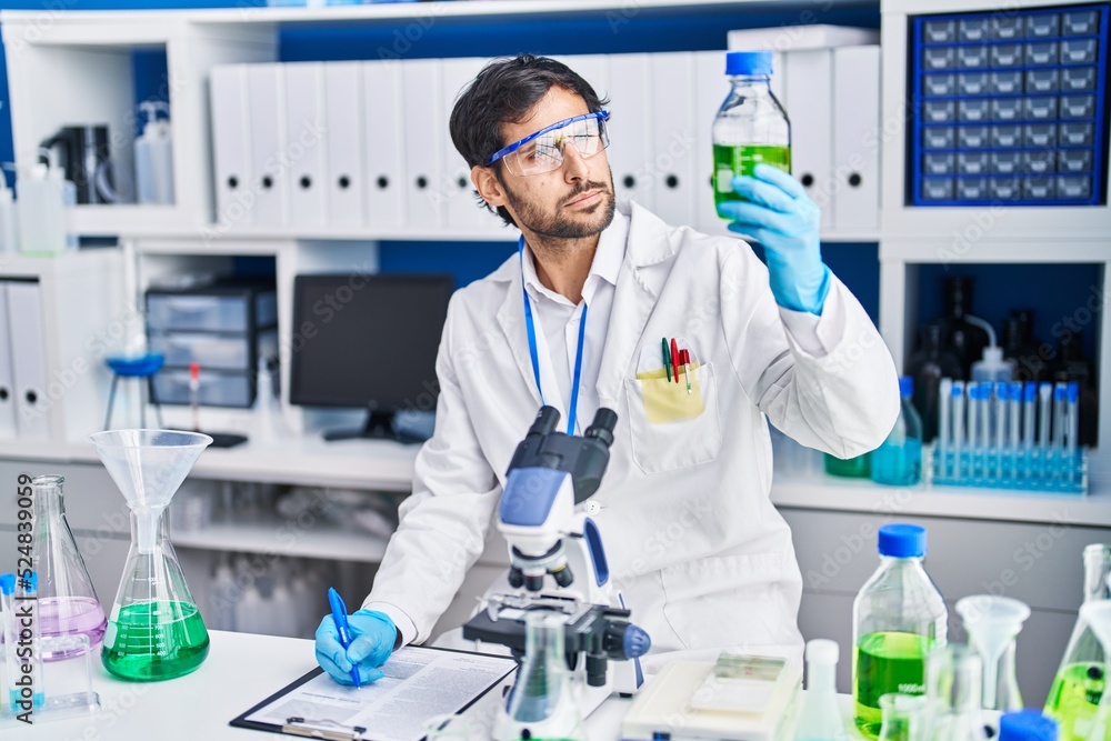 Young hispanic man scientist write on clipboard holding bottle at laboratory Young hispanic man scientist write on clipboard holding bottle at laboratory