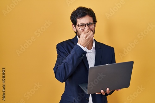 Handsome latin man working using computer laptop smelling something stinky and disgusting, intolerable smell, holding breath with fingers on nose. bad smell