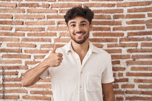 Arab man with beard standing over bricks wall background doing happy thumbs up gesture with hand. approving expression looking at the camera showing success.