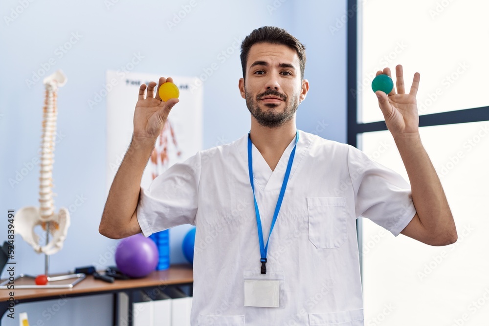 Young hispanic physiotherapist man holding strength balls to train hand muscles relaxed with serious expression on face. simple and natural looking at the camera. Young hispanic physiotherapist man holding strength balls to train hand muscles relaxed with serious expression on face. simple and natural looking at the camera.