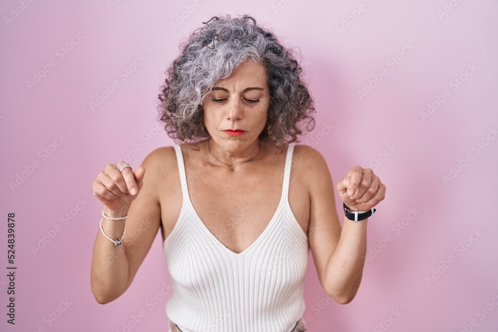 Middle age woman with grey hair standing over pink background pointing down looking sad and upset, indicating direction with fingers, unhappy and depressed. Middle age woman with grey hair standing over pink background pointing down looking sad and upset, indicating direction with fingers, unhappy and depressed.