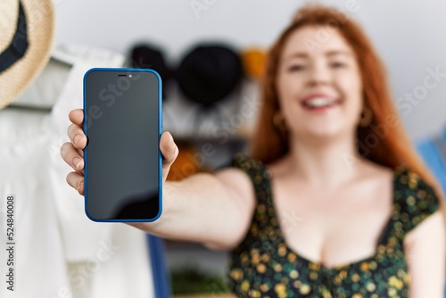 Young redhead woman at retail shop showing smartphone screen looking positive and happy standing and smiling with a confident smile showing teeth