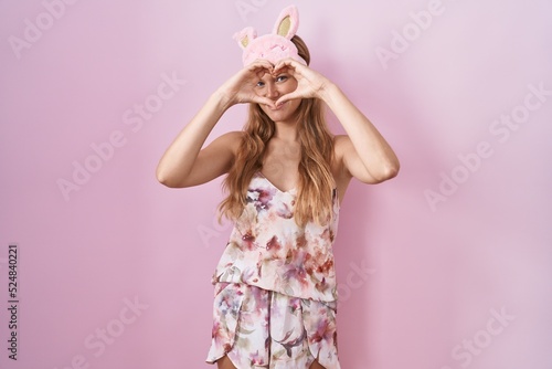 Young caucasian woman wearing sleep mask and pajama doing heart shape with hand and fingers smiling looking through sign