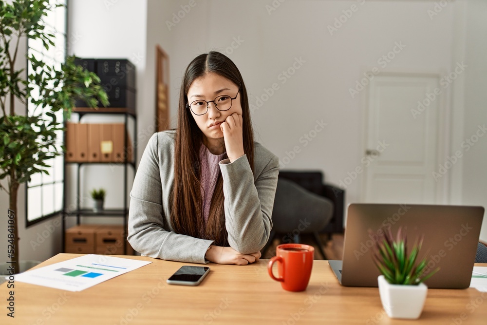 Young chinese business worker wearing business style sitting on desk at office thinking looking tired and bored with depression problems with crossed arms. Young chinese business worker wearing business style sitting on desk at office thinking looking tired and bored with depression problems with crossed arms.