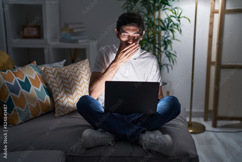 Naklejka premium Young hispanic man using laptop at home at night bored yawning tired covering mouth with hand. restless and sleepiness.