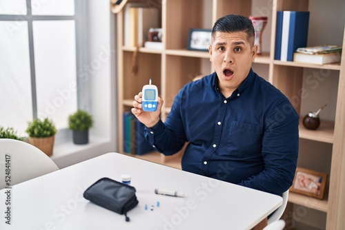 Hispanic young man holding glucometer device scared and amazed with open mouth for surprise, disbelief face