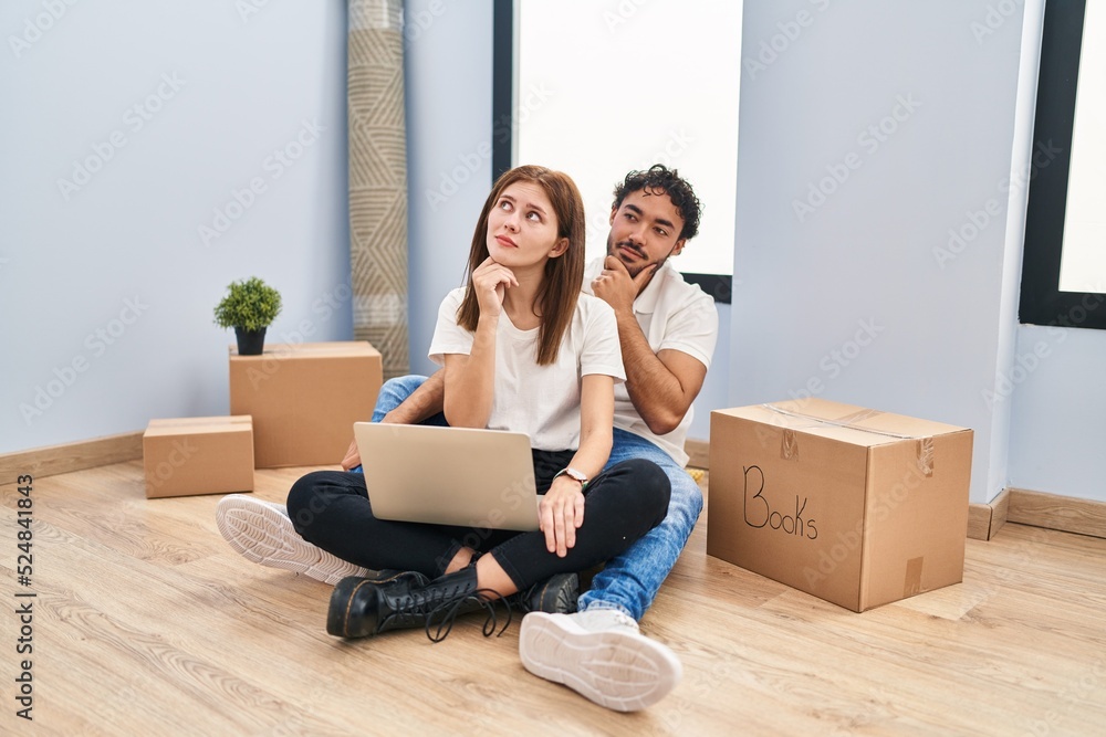 Young couple using laptop at new home with hand on chin thinking about question, pensive expression. smiling with thoughtful face. doubt concept. Young couple using laptop at new home with hand on chin thinking about question, pensive expression. smiling with thoughtful face. doubt concept.