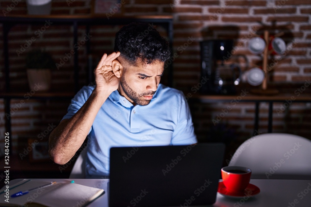 Hispanic man with beard using laptop at night smiling with hand over ear listening an hearing to rumor or gossip. deafness concept. Hispanic man with beard using laptop at night smiling with hand over ear listening an hearing to rumor or gossip. deafness concept.