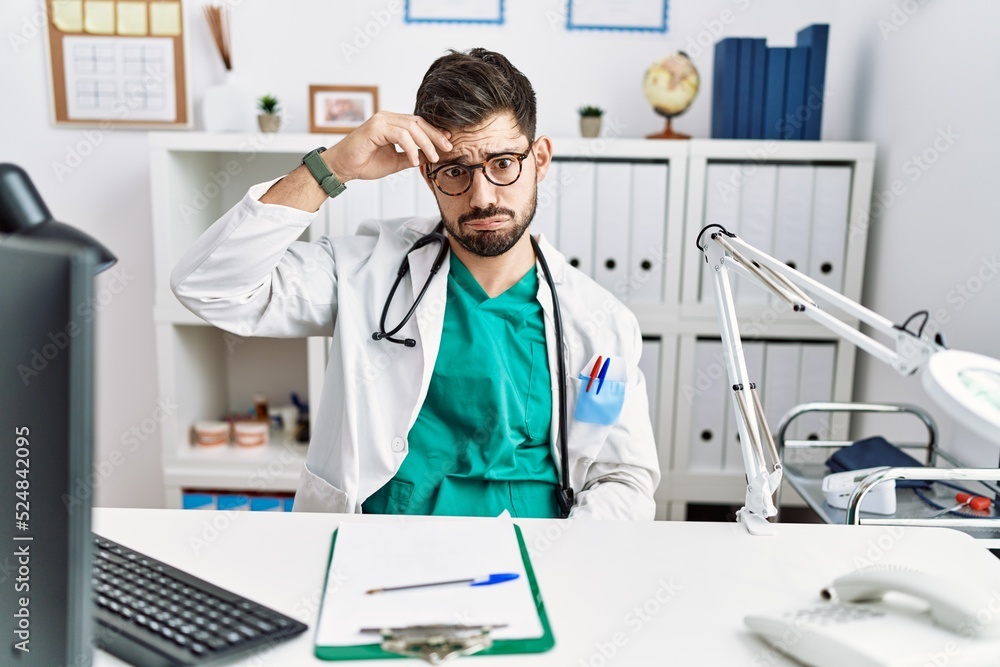 Young man with beard wearing doctor uniform and stethoscope at the clinic worried and stressed about a problem with hand on forehead, nervous and anxious for crisis Young man with beard wearing doctor uniform and stethoscope at the clinic worried and stressed about a problem with hand on forehead, nervous and anxious for crisis