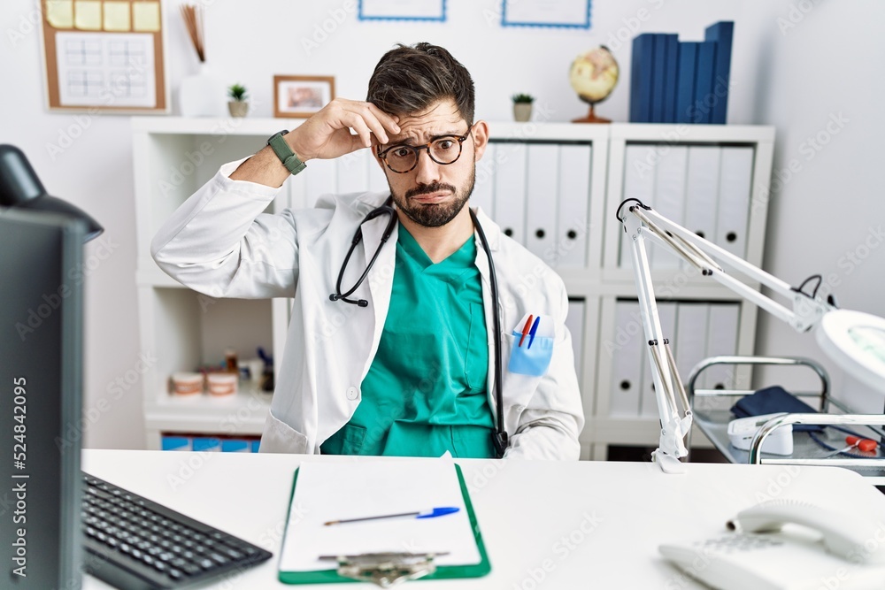 Young man with beard wearing doctor uniform and stethoscope at the clinic worried and stressed about a problem with hand on forehead, nervous and anxious for crisis