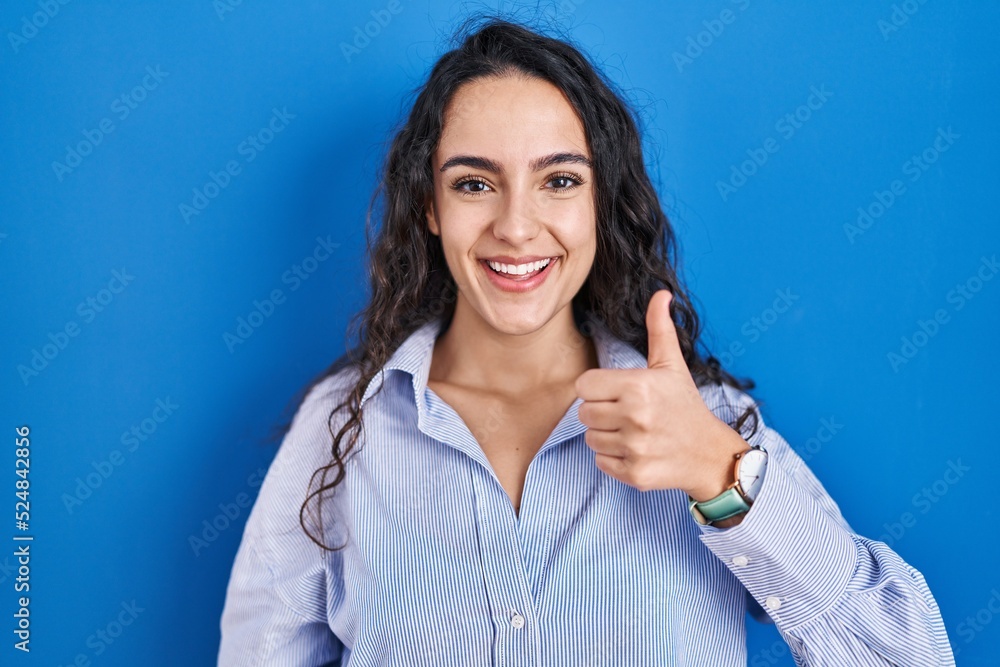 Young brunette woman standing over blue background doing happy thumbs up gesture with hand. approving expression looking at the camera showing success. Young brunette woman standing over blue background doing happy thumbs up gesture with hand. approving expression looking at the camera showing success.