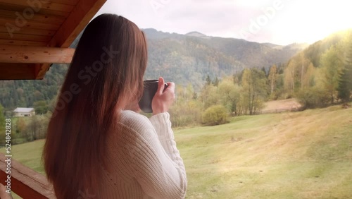 Woman in morning mountains at resort. Unrecognizable woman, back view, drinks coffee, enjoys sunny autumn mountains view. Girl on hotel balcony. Guiet, peaceful vacation in autumn nature concept photo