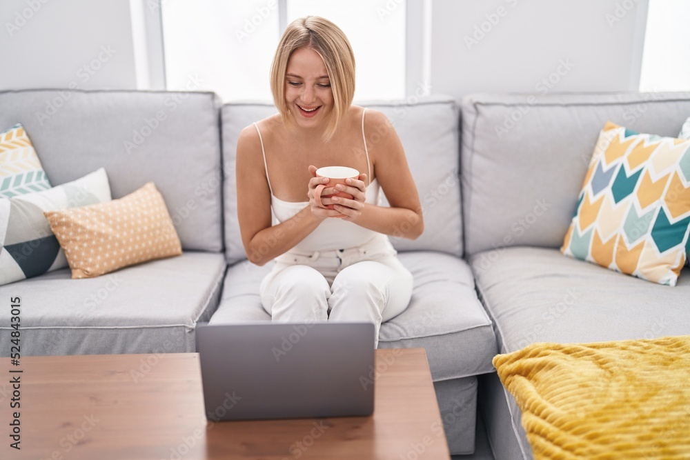Young blonde woman using laptop and drinking coffee sitting on sofa at home Young blonde woman using laptop and drinking coffee sitting on sofa at home