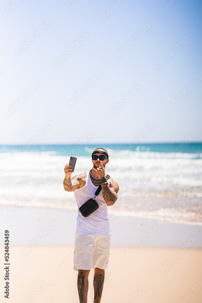 Chico joven musculado posando con cinta en la cabeza en la playa con el smartphone en la mano Chico joven musculado posando con cinta en la cabeza en la playa con el smartphone en la mano