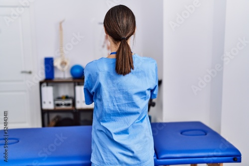 Young hispanic woman wearing physiotherapist uniform standing at clinic standing backwards looking away with crossed arms