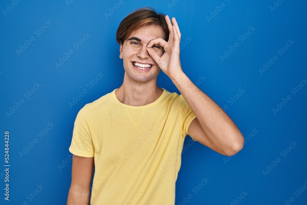 Young man standing over blue background doing ok gesture with hand smiling, eye looking through fingers with happy face. Young man standing over blue background doing ok gesture with hand smiling, eye looking through fingers with happy face.