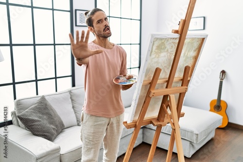 Young hispanic man with beard painting on canvas at home doing stop gesture with hands palms, angry and frustration expression