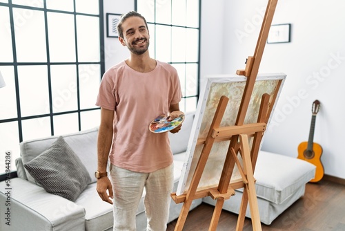 Young hispanic man with beard painting on canvas at home looking away to side with smile on face, natural expression. laughing confident.