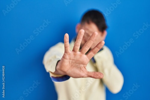 Handsome hispanic man standing over blue background covering eyes with hands and doing stop gesture with sad and fear expression. embarrassed and negative concept.