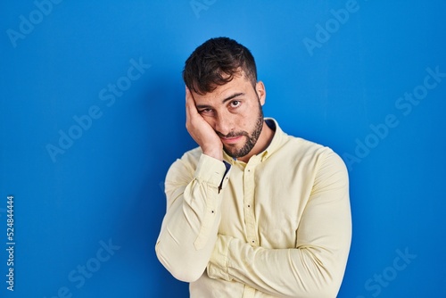 Handsome hispanic man standing over blue background thinking looking tired and bored with depression problems with crossed arms.