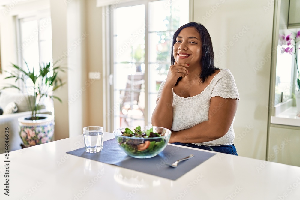 Young hispanic woman eating healthy salad at home looking confident at the camera smiling with crossed arms and hand raised on chin. thinking positive. Young hispanic woman eating healthy salad at home looking confident at the camera smiling with crossed arms and hand raised on chin. thinking positive.