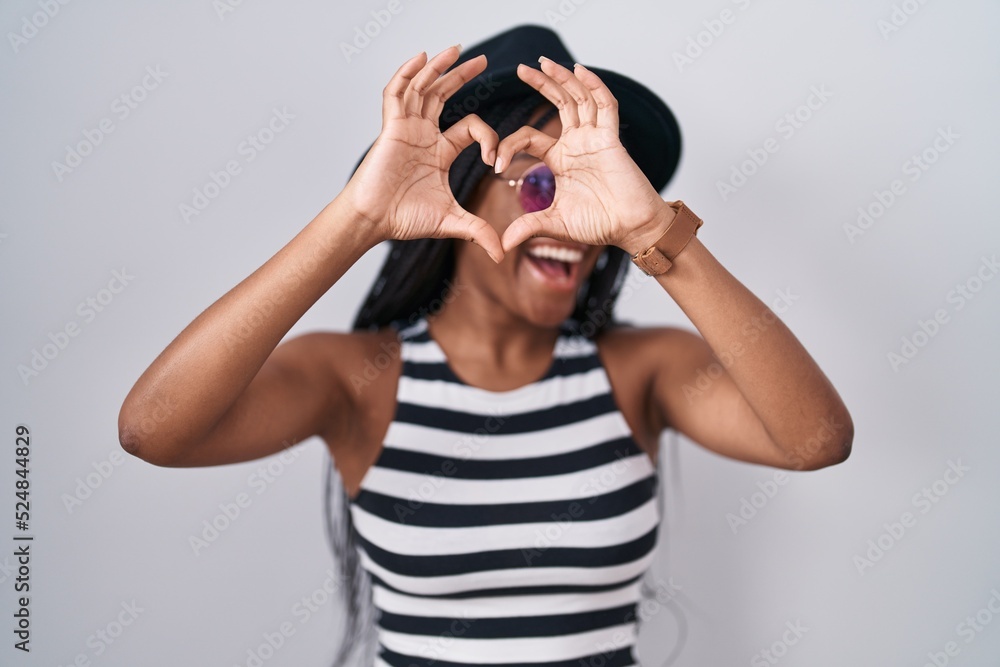 Young african american with braids wearing hat and sunglasses doing heart shape with hand and fingers smiling looking through sign Young african american with braids wearing hat and sunglasses doing heart shape with hand and fingers smiling looking through sign