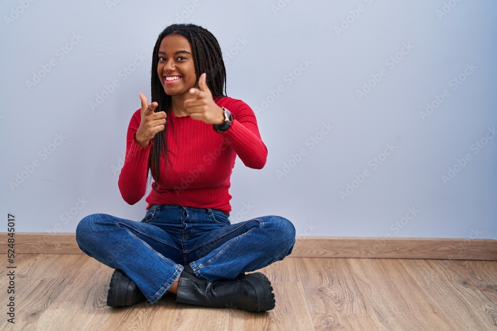 Young african american with braids sitting on the floor at home pointing fingers to camera with happy and funny face. good energy and vibes. Young african american with braids sitting on the floor at home pointing fingers to camera with happy and funny face. good energy and vibes.