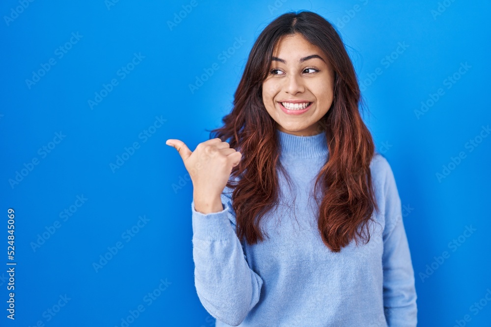 Hispanic young woman standing over blue background smiling with happy face looking and pointing to the side with thumb up. Hispanic young woman standing over blue background smiling with happy face looking and pointing to the side with thumb up.