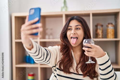 Hispanic young business woman taking a selfie picture drinking a glass of wine sticking tongue out happy with funny expression.