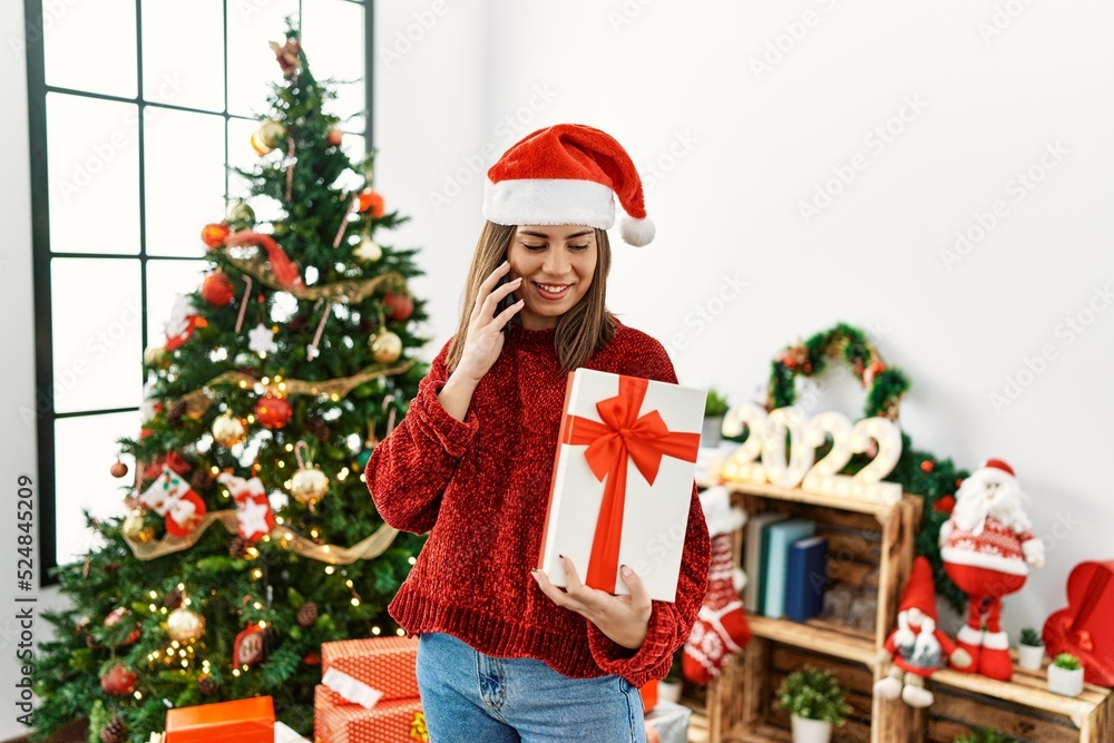 Young hispanic girl talking on the smartphone and holding gift standing by christmas tree at home. Young hispanic girl talking on the smartphone and holding gift standing by christmas tree at home.