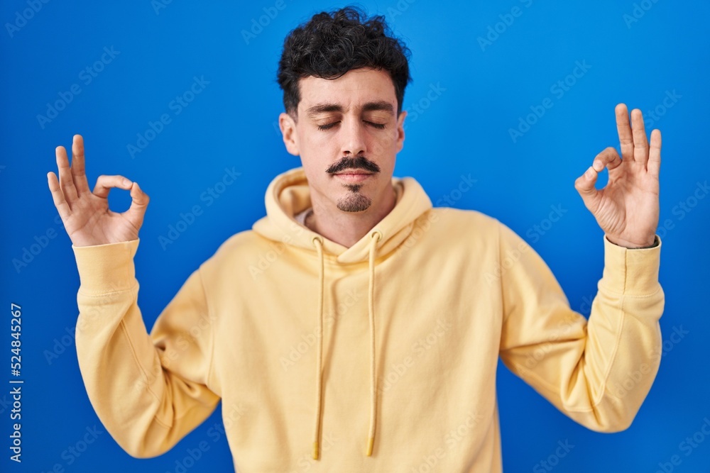 Hispanic man standing over blue background relaxed and smiling with eyes closed doing meditation gesture with fingers. yoga concept. Hispanic man standing over blue background relaxed and smiling with eyes closed doing meditation gesture with fingers. yoga concept.