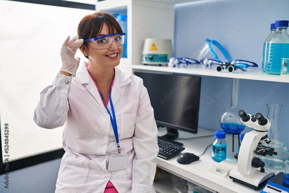Young brunette woman working at scientist laboratory smiling with hand over ear listening an hearing to rumor or gossip. deafness concept. Young brunette woman working at scientist laboratory smiling with hand over ear listening an hearing to rumor or gossip. deafness concept.
