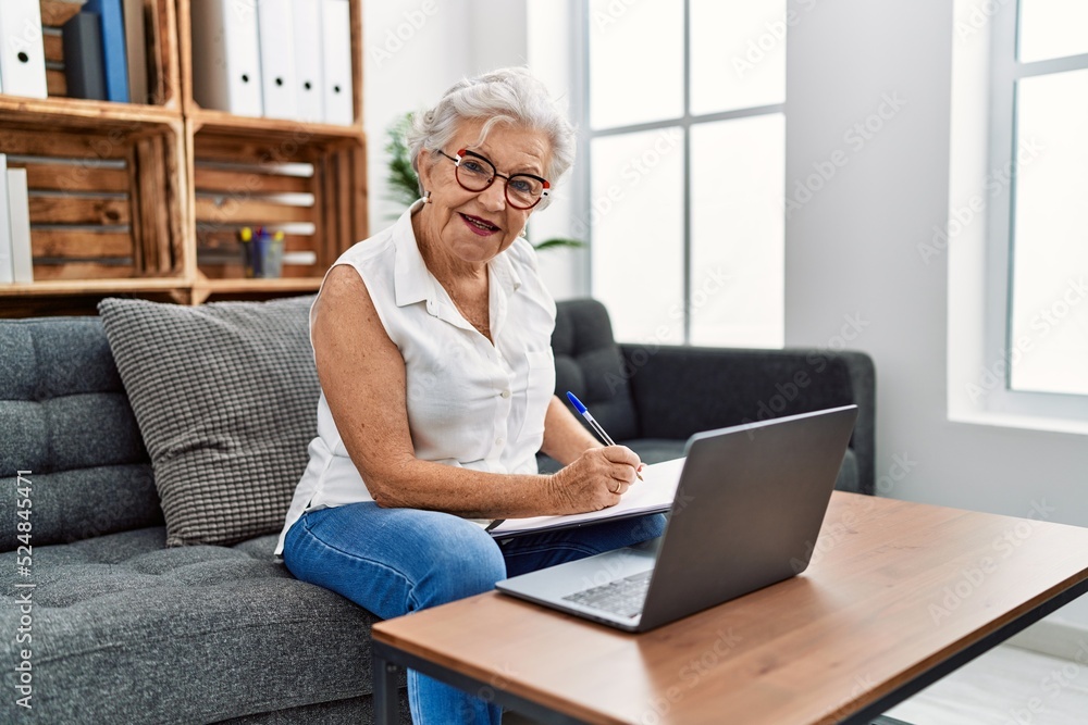 Senior grey-haired woman psychologist writing on clipboard using laptop at psychology center Senior grey-haired woman psychologist writing on clipboard using laptop at psychology center