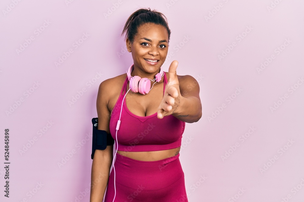 Young african american girl wearing gym clothes and using headphones smiling friendly offering handshake as greeting and welcoming. successful business.