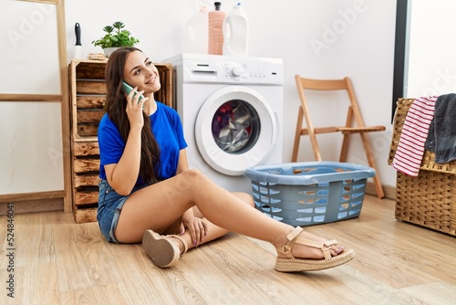 Young caucasian woman talking on the smartphone waiting for washing machine at laundry room