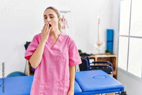 Young blonde woman working at pain recovery clinic bored yawning tired covering mouth with hand. restless and sleepiness.