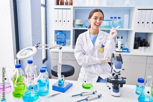 Young brunette woman working at scientist laboratory smiling happy pointing with hand and finger to the side