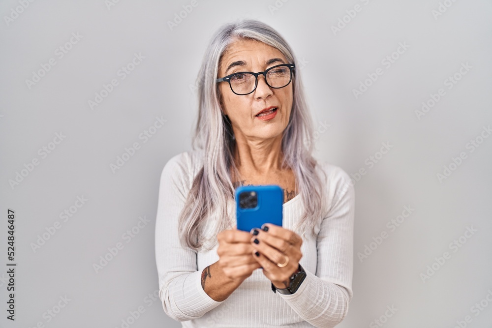 Middle age woman with grey hair using smartphone typing message winking looking at the camera with sexy expression, cheerful and happy face.
