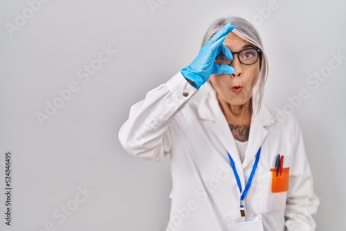 Middle age woman with grey hair wearing scientist robe doing ok gesture shocked with surprised face, eye looking through fingers. unbelieving expression.