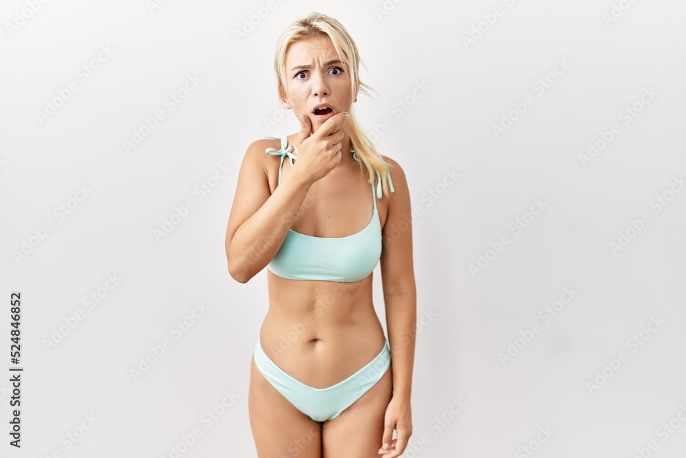 Fototapeta premium Young caucasian woman wearing bikini over isolated background looking fascinated with disbelief, surprise and amazed expression with hands on chin