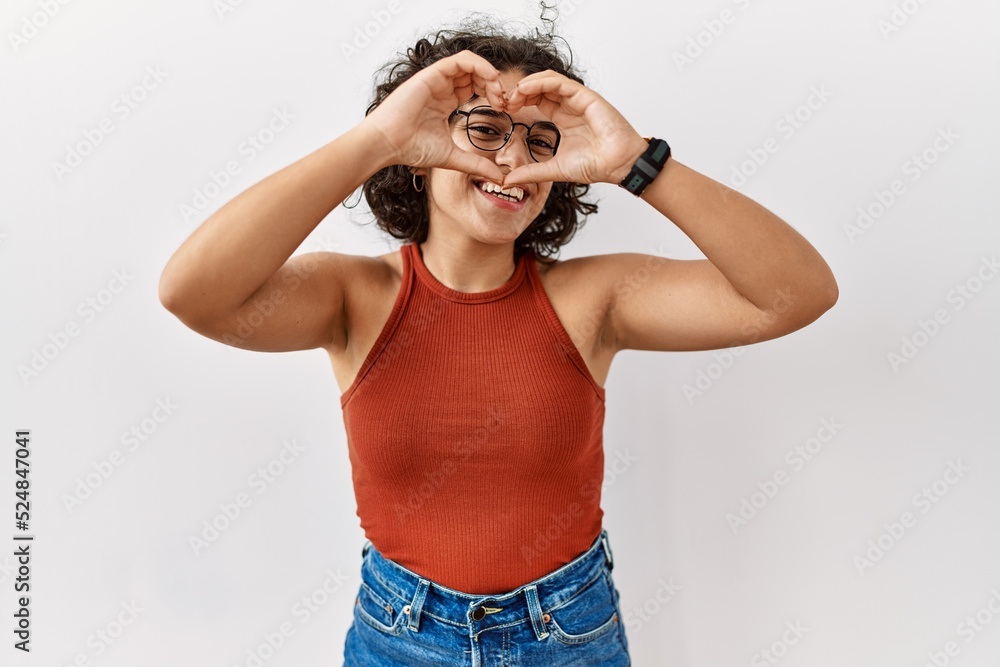 Young hispanic woman wearing glasses standing over isolated background doing heart shape with hand and fingers smiling looking through sign Young hispanic woman wearing glasses standing over isolated background doing heart shape with hand and fingers smiling looking through sign